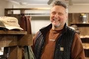 Nisjebutikk med mann i brun crewneck med brodert "Bybonde" på i hvitt, beige/naturfaget vintage caps med brun brodering på hyll.
